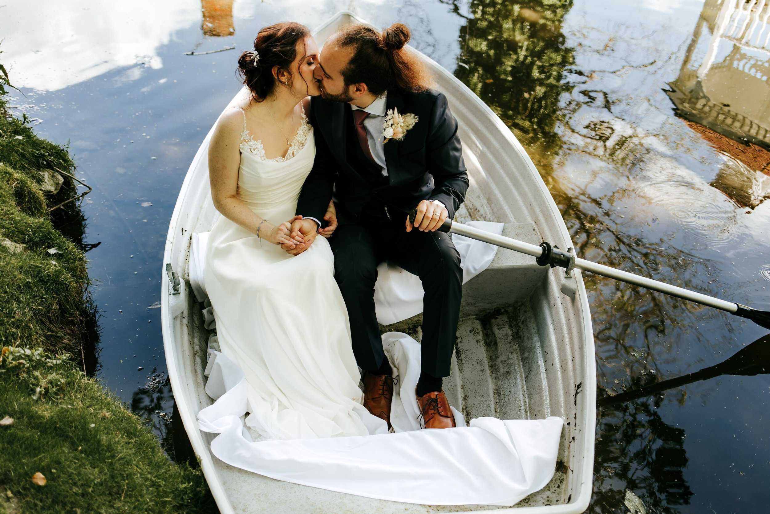 A bride and groom share a kiss while sitting in a small rowing boat on a tranquil body of water, surrounded by greenery.