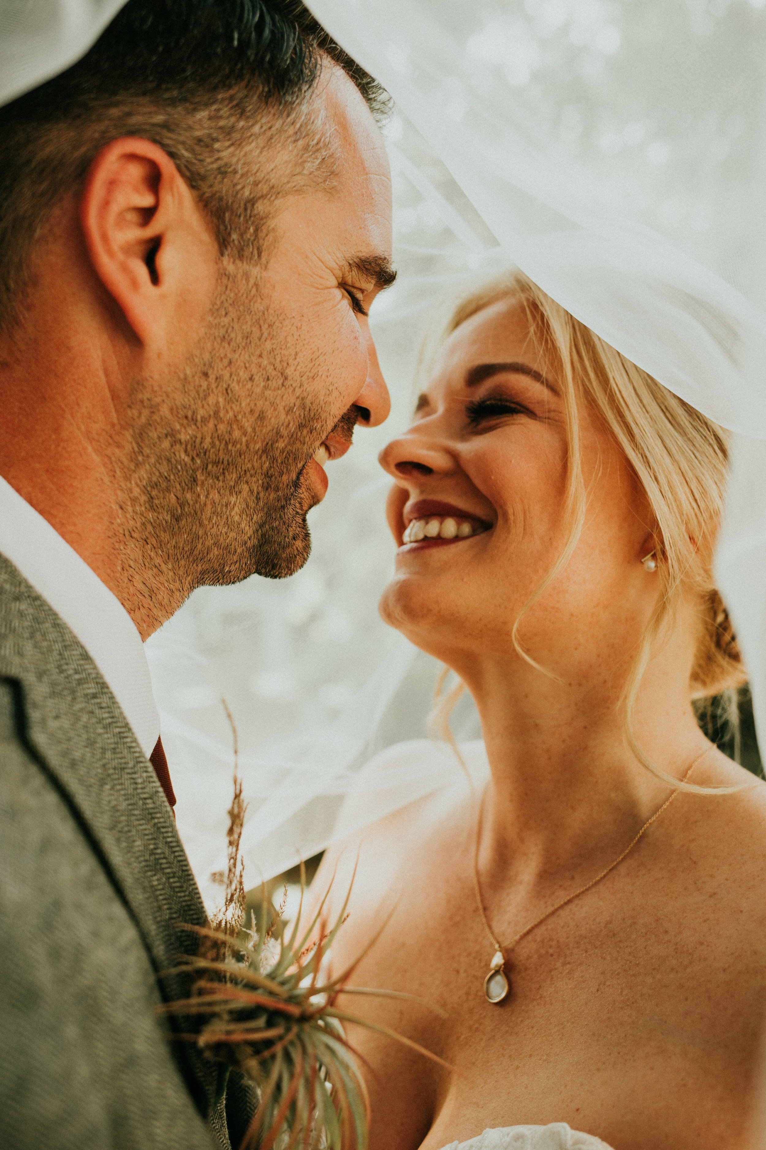 A close-up photograph of a smiling couple sharing an intimate moment under a veil, with soft natural lighting enhancing their joyful expressions.