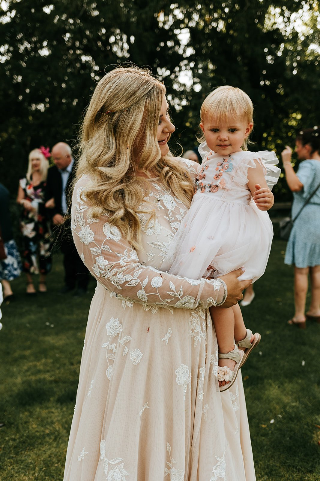 A bride in a floral embroidered wedding dress holds a small girl in a white dress, both smiling in a sunny outdoor setting with guests mingling in the background.