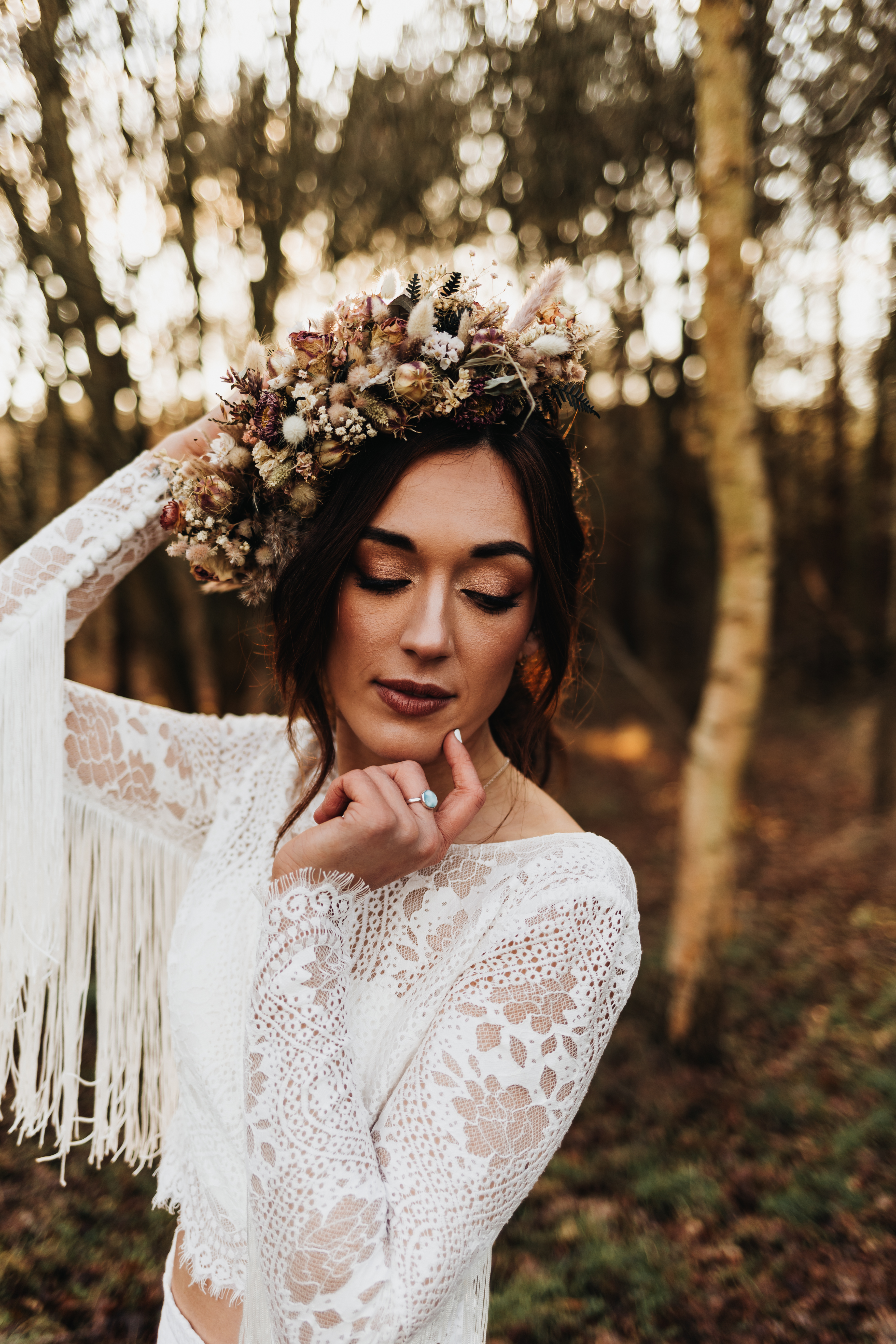 A woman in a white lace top with fringe gently touches her face, wearing a floral crown, set in a wooded area with soft sunlight filtering through the trees.