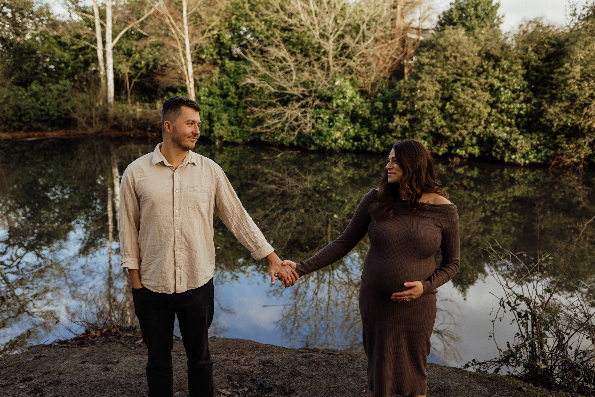 Pregnant woman and man holding hands and looking at each other by a lake with trees in the background during a maternity photoshoot in Suffolk