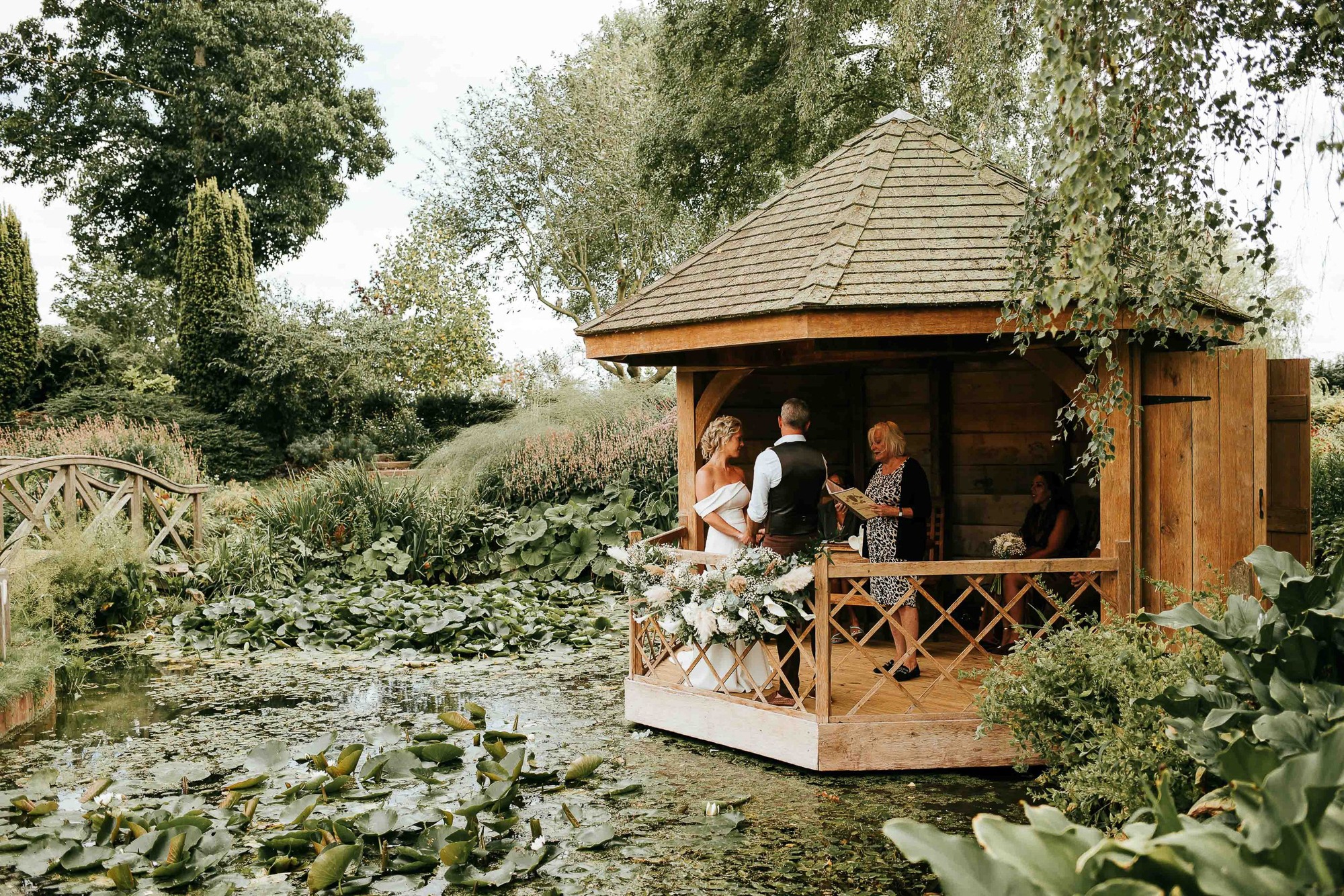 Couple in wedding attire standing inside a wooden gazebo by a pond with lily pads, surrounded by lush greenery and an officiant reading from a book.